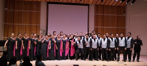 Large choir group standing on stage wearing black and pink formal attire with projection screen in background